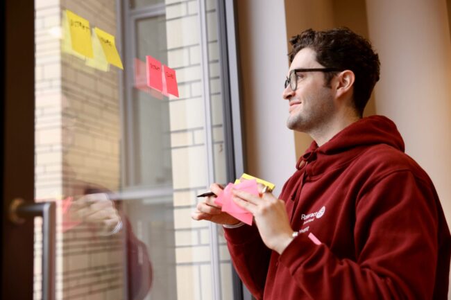 Mann mit dunkelroter Sweatjacke klebt Post-Its an ein Fenster.