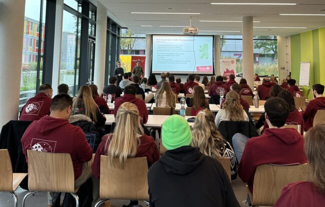 Blick von hinten auf das voll besetzte Plenum. Vor den sitzenden Menschen wird ein Vortrag gehalten mit einer Präsentation, die über einen Beamer ausgespielt wird.