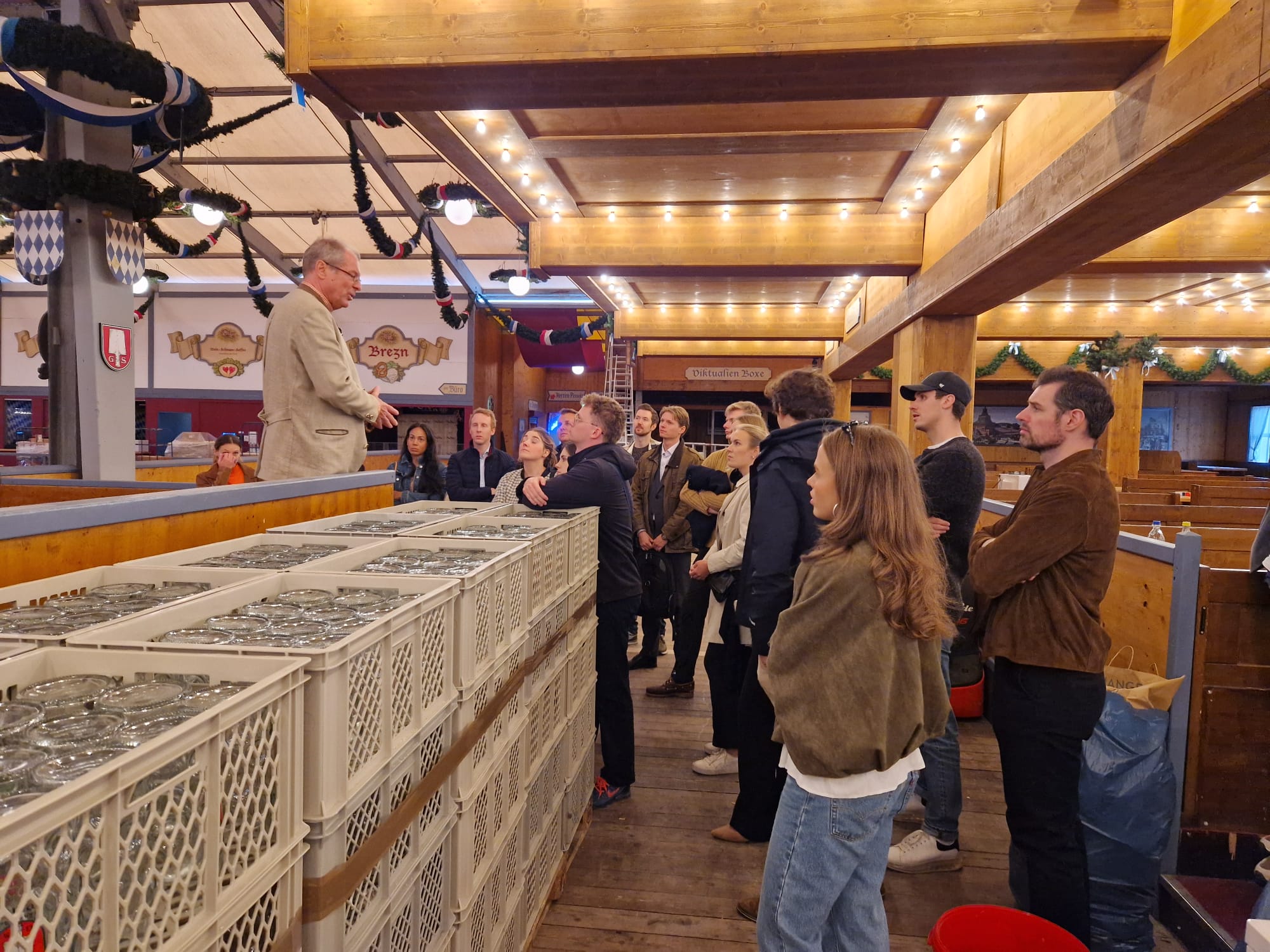 Eine Gruppe junger Erwachsener steht in einem leeren Wiesn-Zelt und blickt nach links zu einem Mann mittleren Alters.
