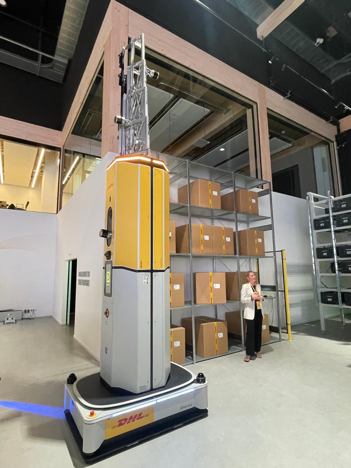Frau steht in einer hohen Lagerhalle und blickt auf einen DHL-Roboter.